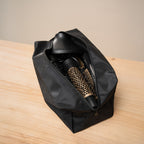 Trousse de rangement noire ouverte sur un plan de travail en bois clair, laissant voir la brosse ronde dorée et les autres embouts rangés à l'intérieur