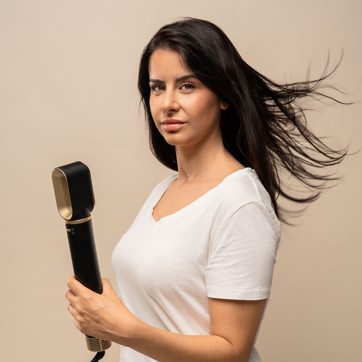 Femme brune en t-shirt blanc tenant l'appareil de coiffage noir et doré, cheveux soulevés par le souffle, fond beige uni