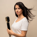 Femme brune en t-shirt blanc tenant l'appareil de coiffage noir et doré, cheveux soulevés par le souffle, fond beige uni