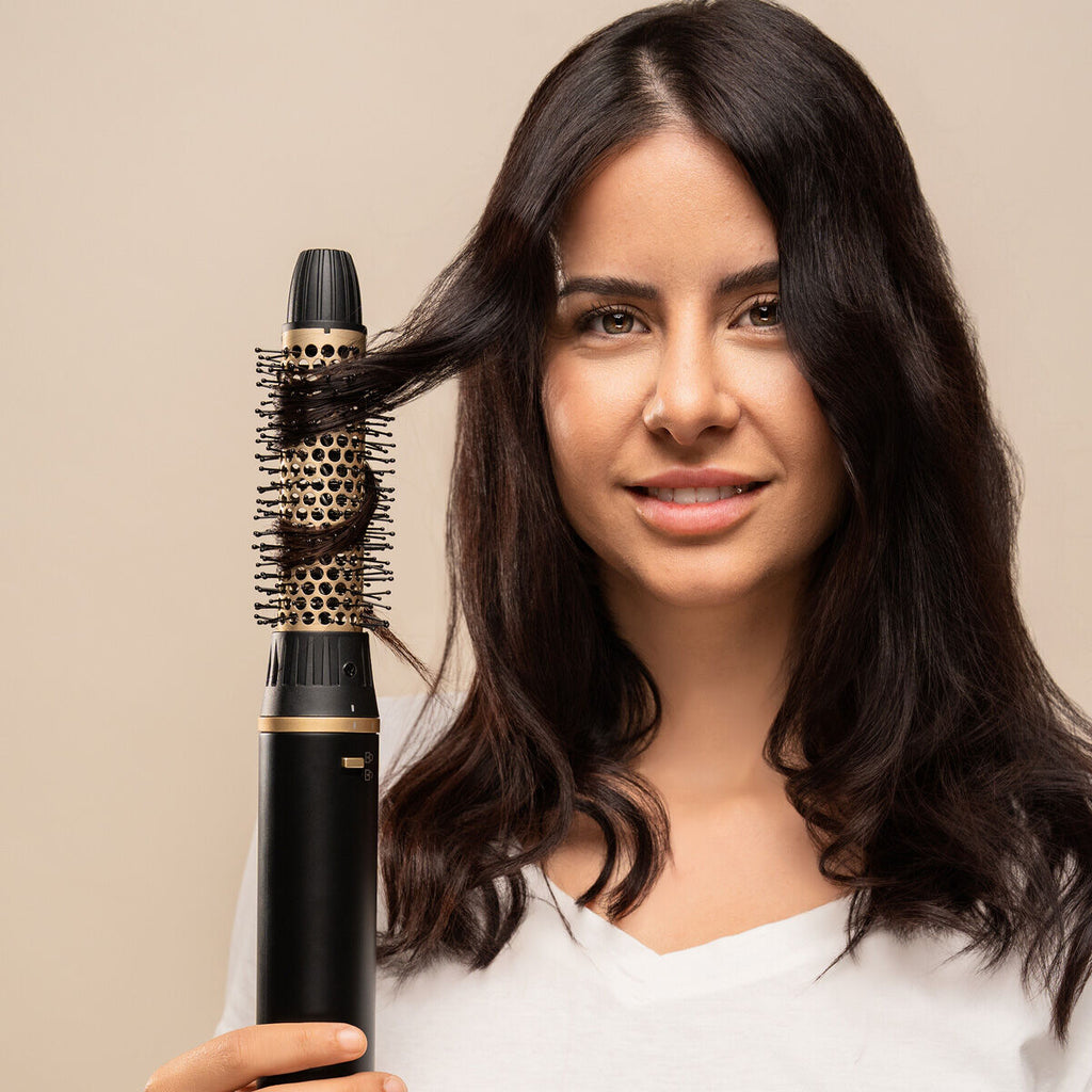 Femme brune souriante tenant l'appareil équipé de sa brosse ronde dorée, une mèche enroulée autour de la brosse, fond beige