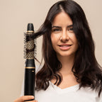 Femme brune souriante tenant l'appareil équipé de sa brosse ronde dorée, une mèche enroulée autour de la brosse, fond beige