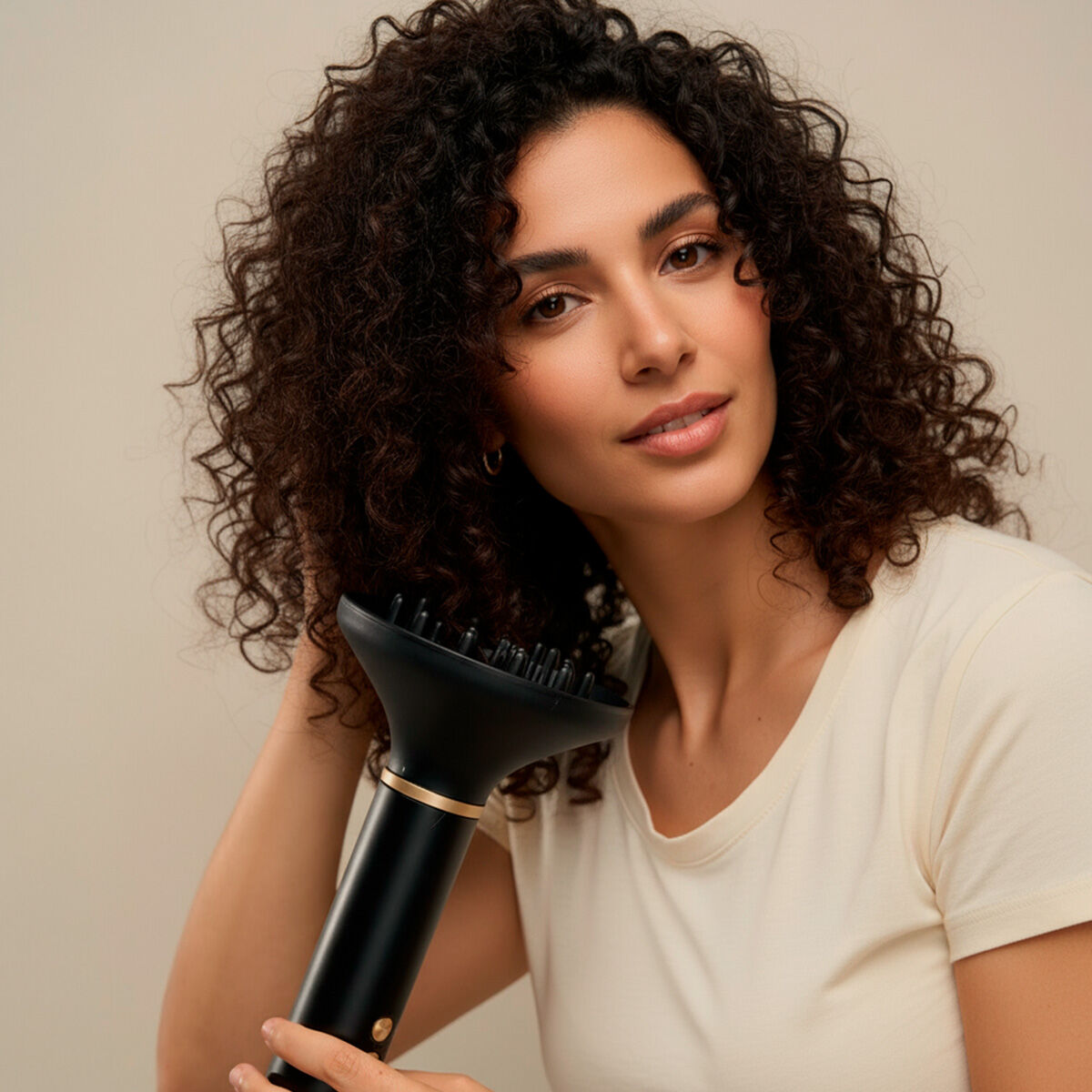 Femme aux cheveux bouclés tenant l'appareil muni de son diffuseur noir, t-shirt écru, fond beige
