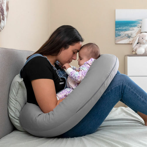 Mère assise sur un lit, front contre front avec son bébé installé dans le coussin d'allaitement gris